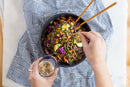 A bowl of Asian style noodles with chopsticks and a person putting garlic salt atop it on a blue striped cloth.