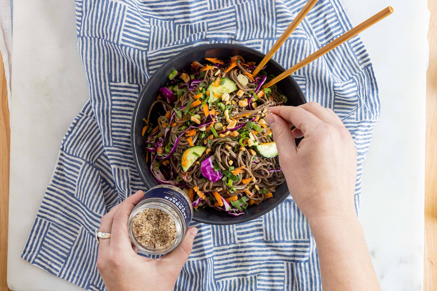 A bowl of Asian style noodles with chopsticks and a person putting garlic salt atop it on a blue striped cloth.