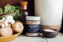 A jar of Jacobson Salt Co. black pepper salt next to a bowl of salt and a bowl of produce on a marble surface.