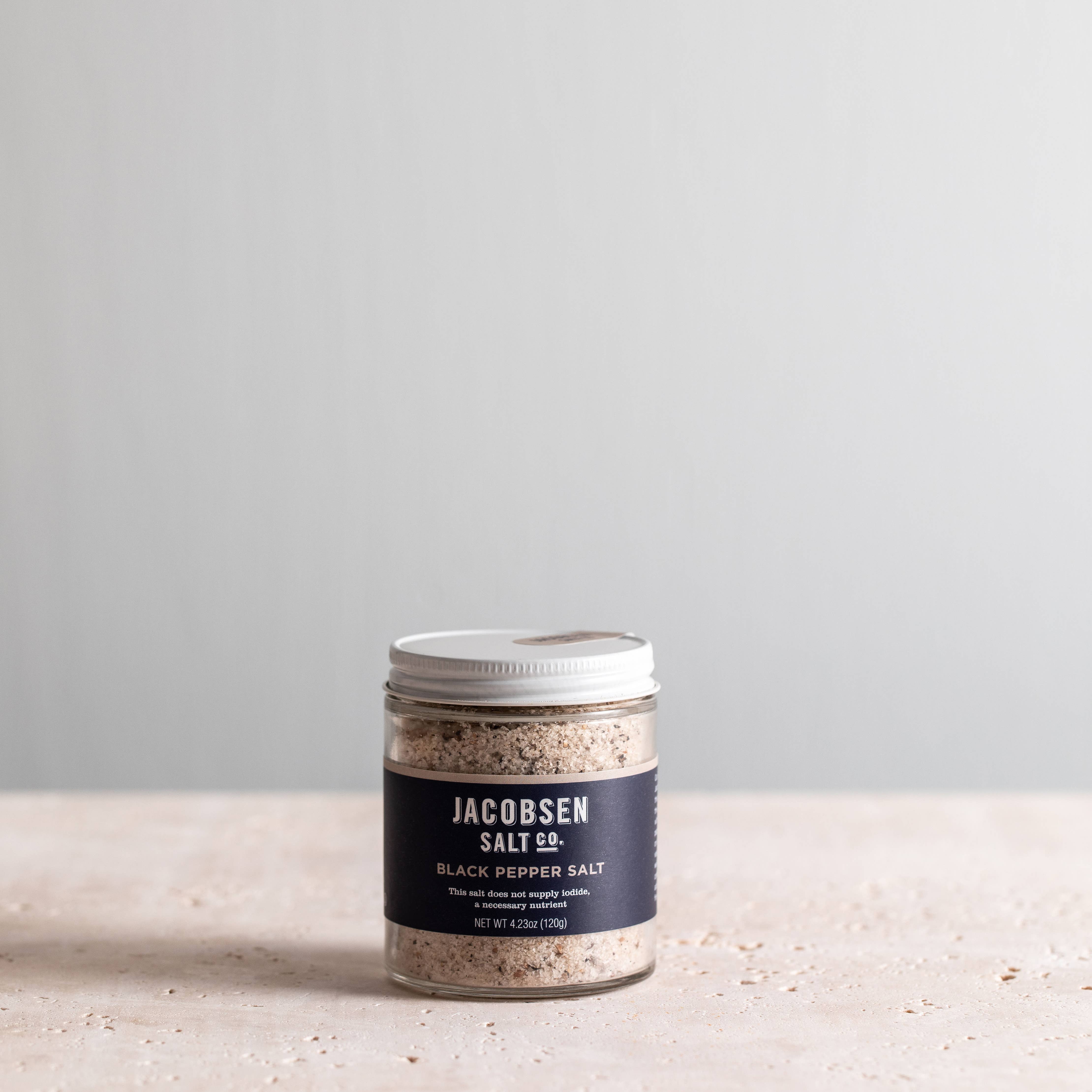A clear glass jar with a silver lid containing black pepper salt, labeled 'JACOBSEN SALT CO. BLACK PEPPER SALT' on a simple background.