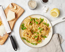 A pan of pasta with seasonings on a marble surface with half a lemon, cutting board with cheese and garlic and a grater, and salt.