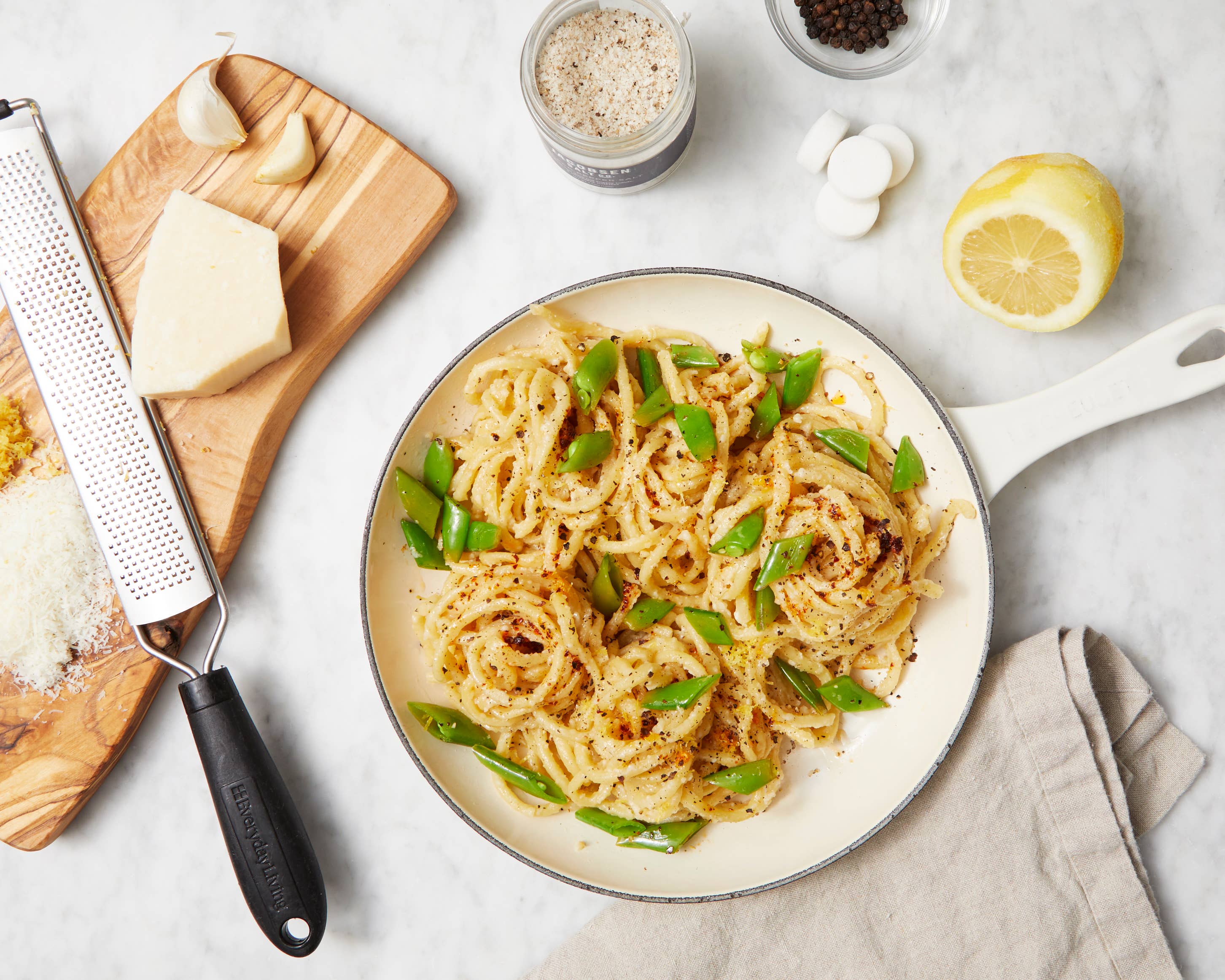 A pan of pasta with seasonings on a marble surface with half a lemon, cutting board with cheese and garlic and a grater, and salt.