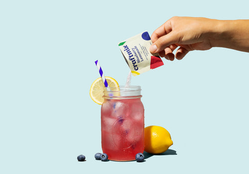 A person pouring a single packet of craftmix into a mason jar full of pink liquid with lemons and blueberries around on a pale blue background.