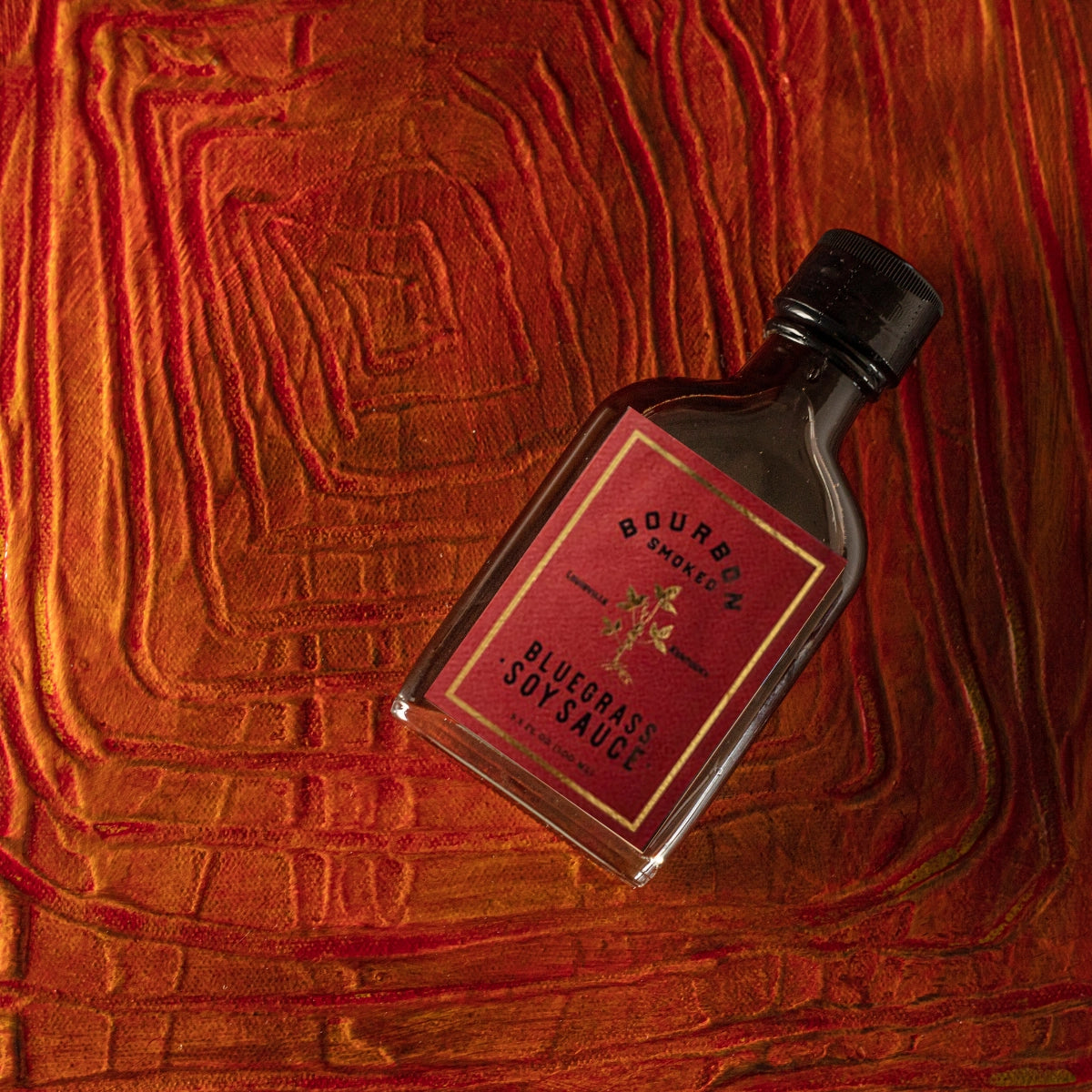 Bottle of Bourbon Smoked Bluegrass Soy Sauce on a textured red wood background
