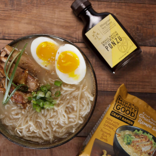 Bowl of ramen with a soft-boiled egg, green onions, and a bottle of ponzu sauce on a wooden surface.