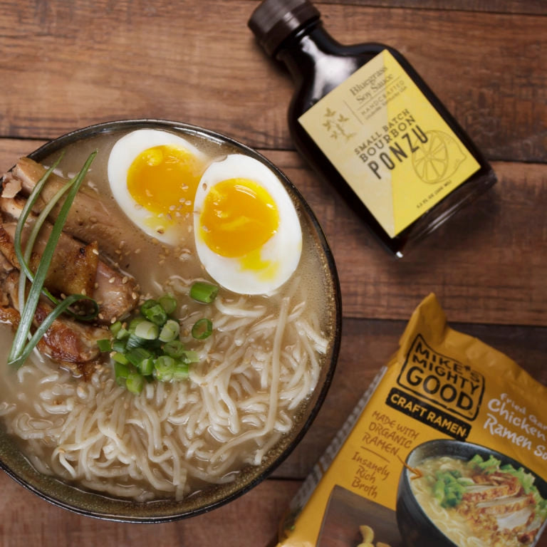 Bowl of ramen with a soft-boiled egg, green onions, and a bottle of ponzu sauce on a wooden surface.