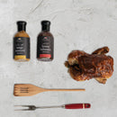 Two bottles of BBQ sauce with a cooked BBQ chicken and cooking utensils on a neutral background.