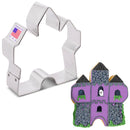 A castle shaped cookie cutter with a sticker that says 'made in USA' next to an example of a finished cookie made to look like a purple haunted house with a small ghost in a window.