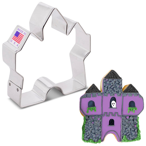 A castle shaped cookie cutter with a sticker that says 'made in USA' next to an example of a finished cookie made to look like a purple haunted house with a small ghost in a window.