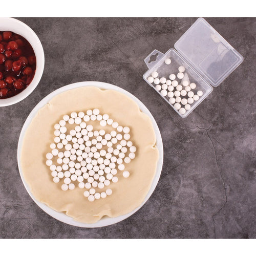 Pie crust with white pie weights on a gray surface, next to a bowl of red berries and a container of additional pie weights.