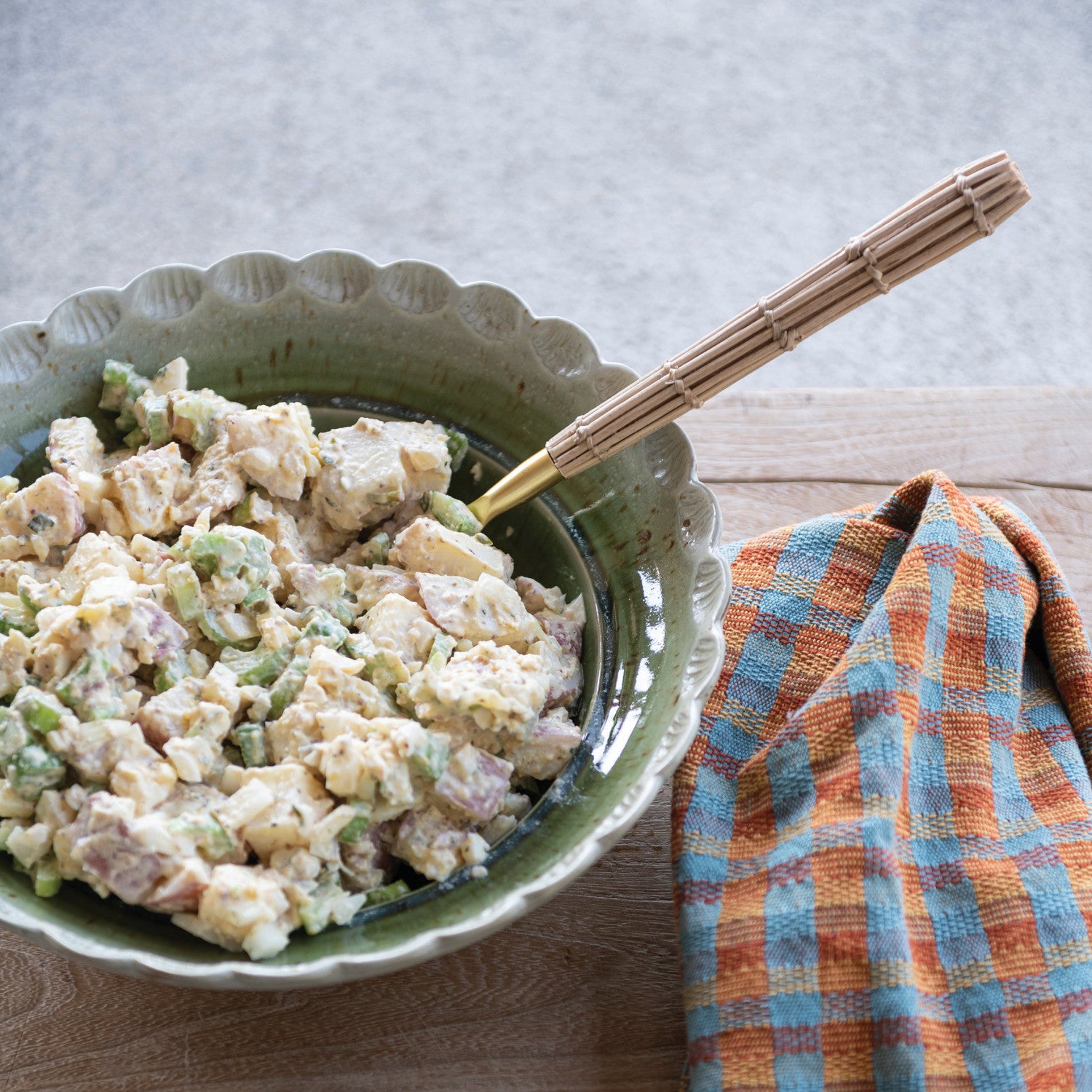 Green bowl of potato salad with a wooden spoon on a wooden surface with a plaid towel.