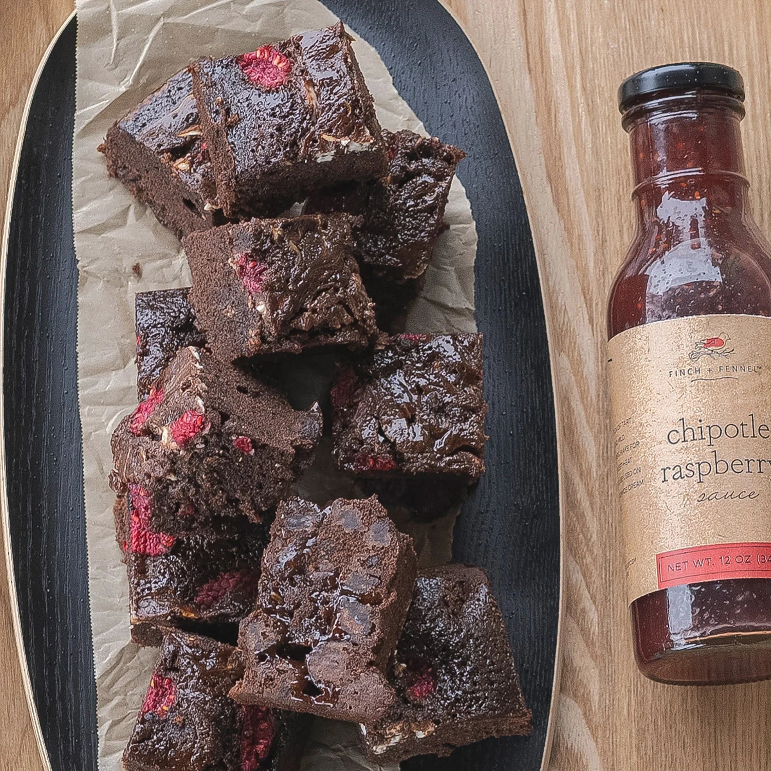 Chocolate brownies with raspberry pieces on a plate next to a bottle of chipotle raspberry sauce.
