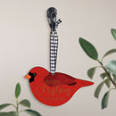 A wooden cardinal ornament that can be customized, hanging from a checkered ribbon with a plant in the background.