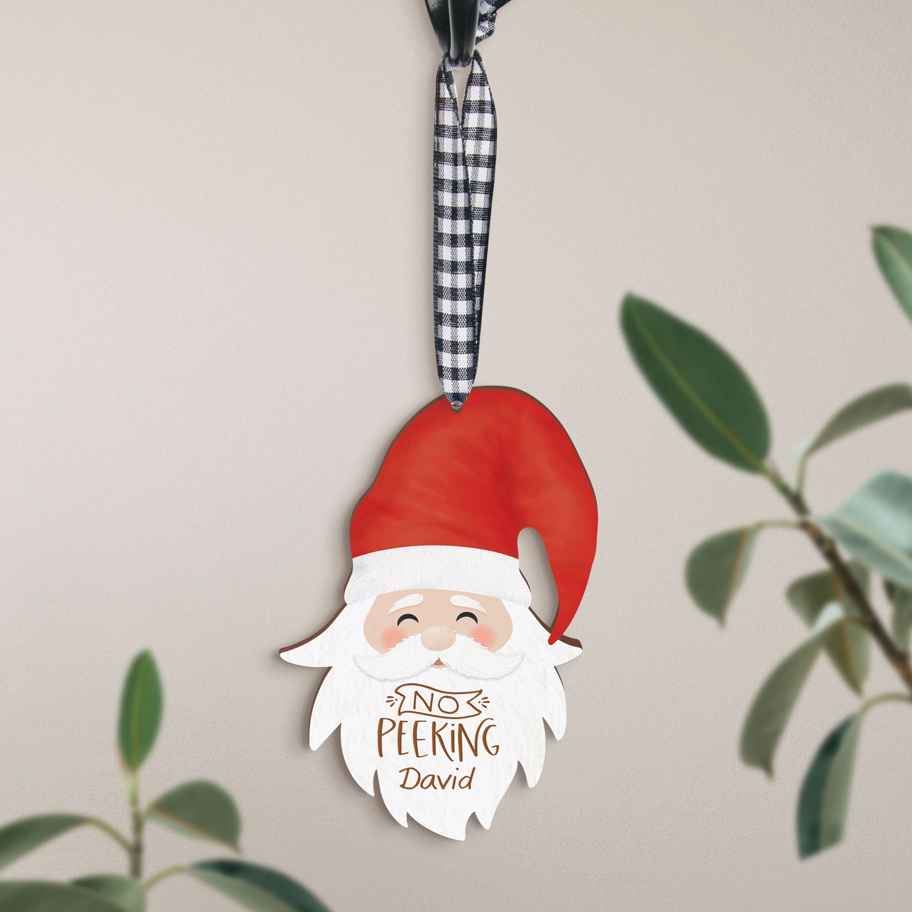 A wooden Santa ornament that can be customized, hanging from a checkered ribbon with a plant in the background.