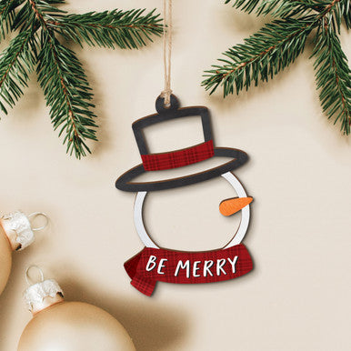 Decorative customizable snowman ornament with 'Be Merry' text hanging among Christmas tree branches.