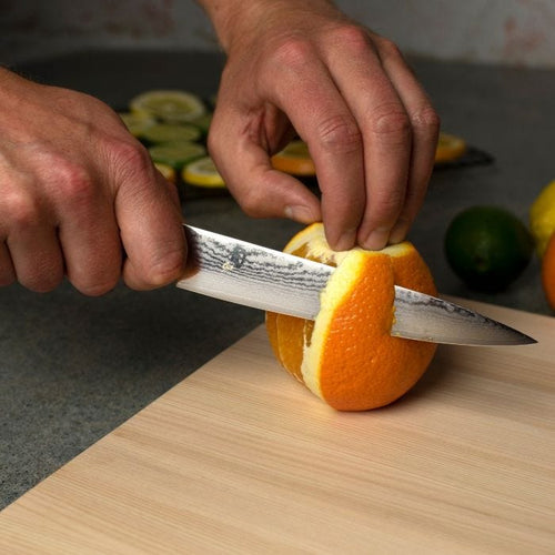 Person cutting an orange with a knife on a wooden cutting board.