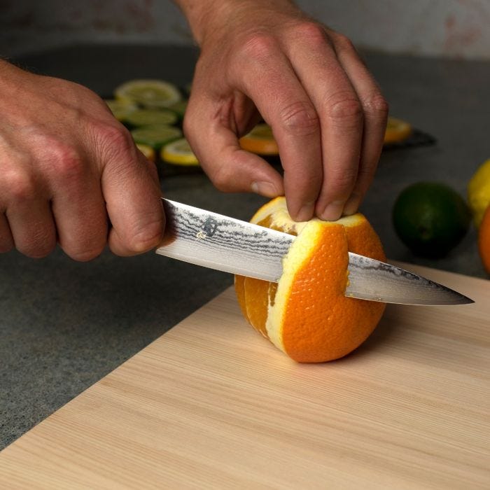 Person cutting an orange with a knife on a wooden cutting board.