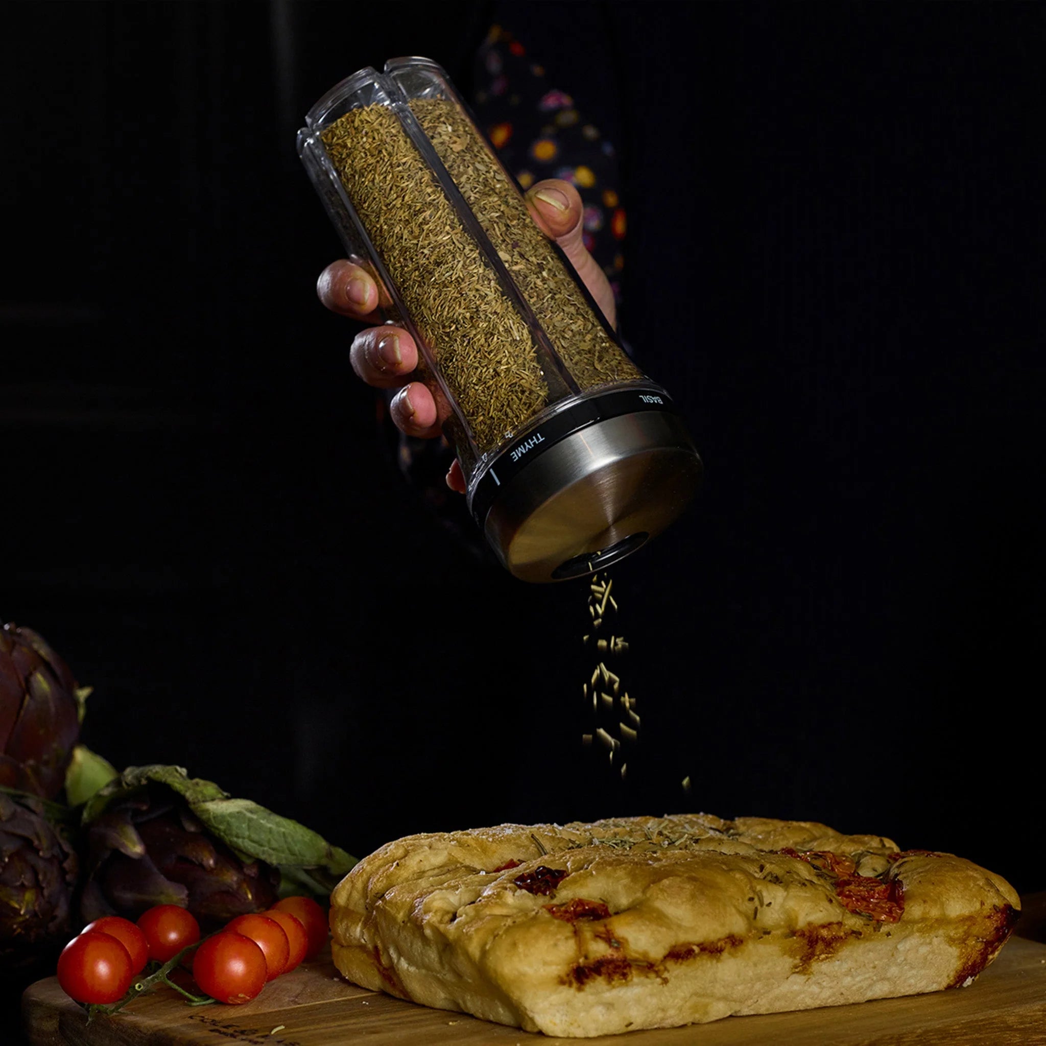 Hand holding a spice shaker over a loaf of bread with vegetables on a dark background
