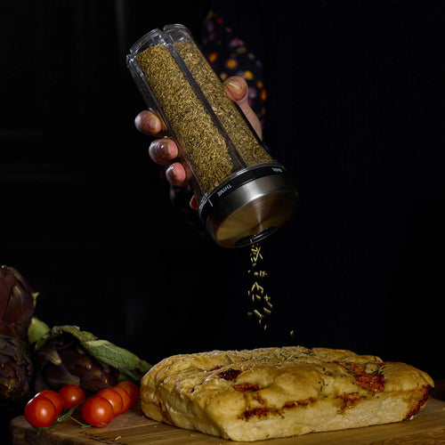 Hand holding a spice shaker over a loaf of bread with vegetables on a dark background