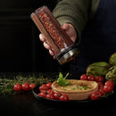 Hand holding a spice shaker over a dish with vegetables on a dark background