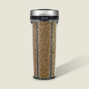Cole & Mason oregano herb grinder with a clear cylindrical body showing the dried green-brown oregano inside, a black band with white text, and a brushed silver metal lid, set against a solid light beige background.
