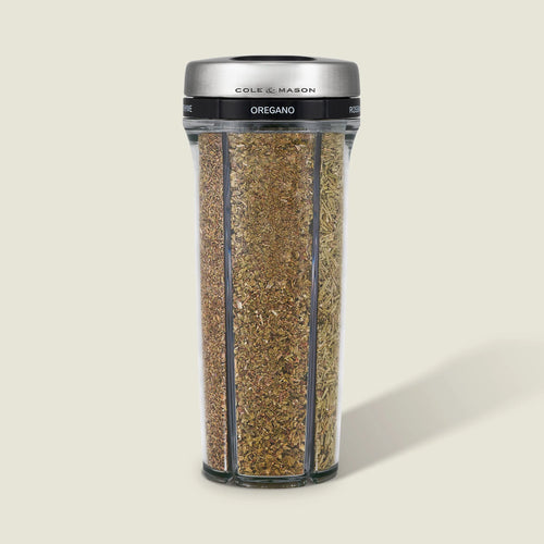 Cole & Mason oregano herb grinder with a clear cylindrical body showing the dried green-brown oregano inside, a black band with white text, and a brushed silver metal lid, set against a solid light beige background.