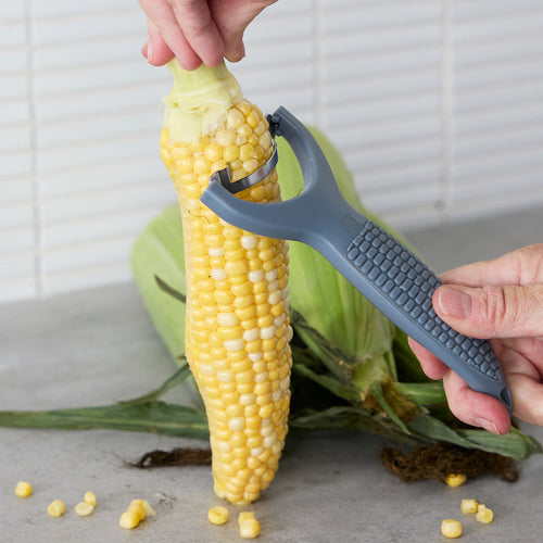 A person using a stainless steel corn stripper on an ear of corn, with more corn on the table nearby.
