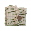 A folded cotton apron with aligator print, accompanied by a chef hat and oven mitt, tied together with a ribbon and a tag.