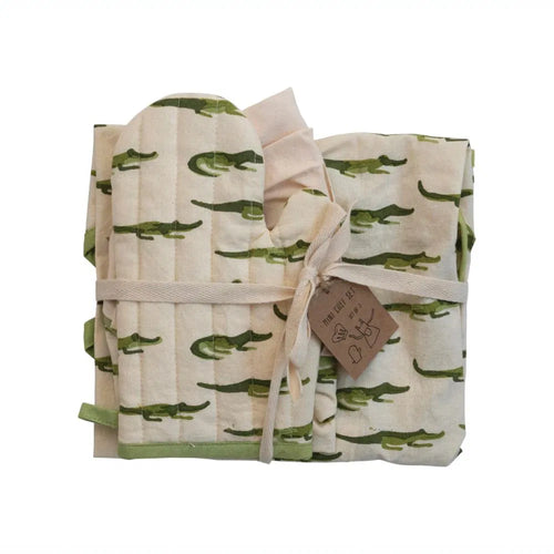 A folded cotton apron with aligator print, accompanied by a chef hat and oven mitt, tied together with a ribbon and a tag.