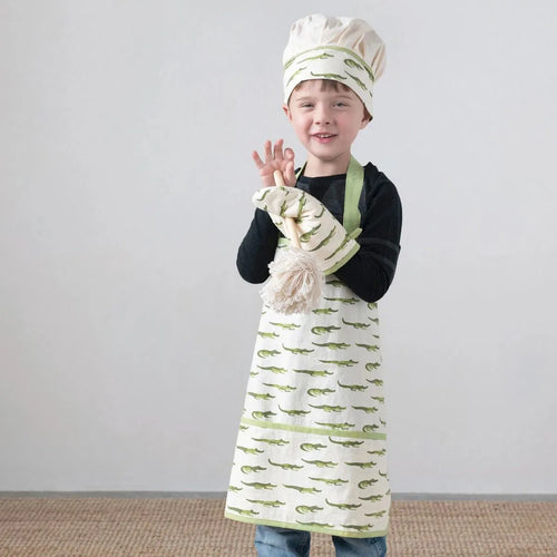 Child wearing an alligator-patterned apron, oven mitt, and chef's hat on a plain background