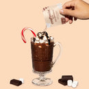 Craftmix hot chocolate being poured into a glass with a candy cane, surrounded by chocolate pieces and marshmallows on a beige background.
