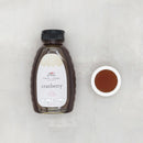 A bottle of Finch + Fennel Cranberry Honey with a small bowl of honey next to it on a gray background.