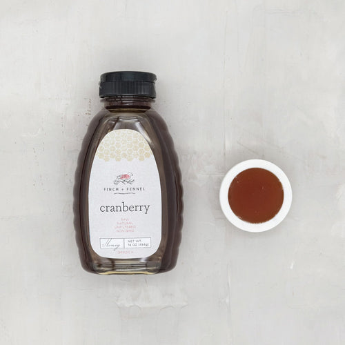 A bottle of Finch + Fennel Cranberry Honey with a small bowl of honey next to it on a gray background.