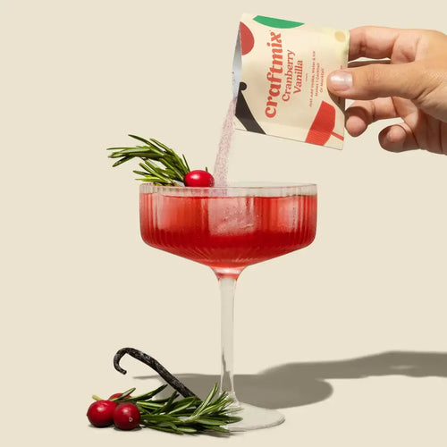 Hand pouring a craftmix Cranberry Vanilla packet into a red cocktail with garnishes on a beige background