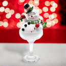 A glass snowman ornament with a happy facial expression, wearing a hat and a coat, holding a candy cane, set against a blurred red and white Christmas-themed backdrop.