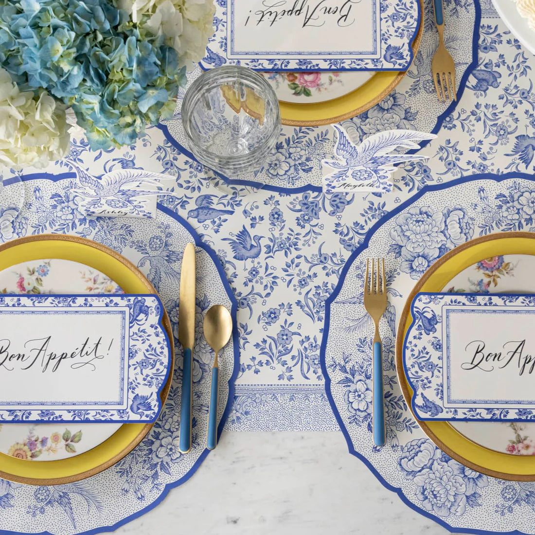 Place settings featuring paper placemats with pheasants and florals in blue and white with blue and gold flatware.