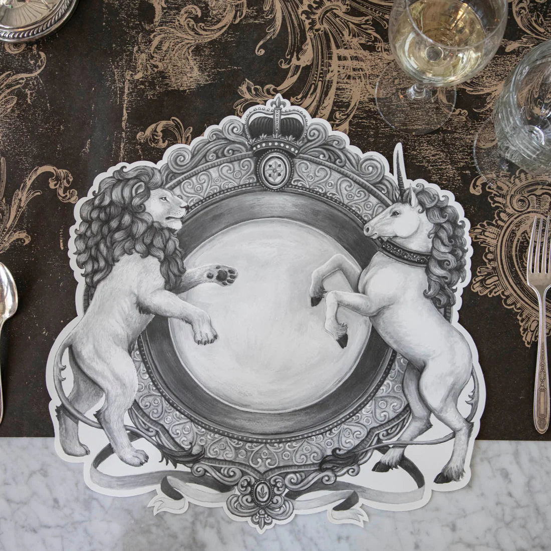 A black and white placemat with a regal crest style illustration featuring a lion on one side and a unicorn on the other resting on a decorative brown table runner with drinking glasses.