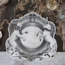 A black and white placemat with a regal crest style illustration featuring a lion on one side and a unicorn on the other resting on a decorative brown table runner with drinking glasses.