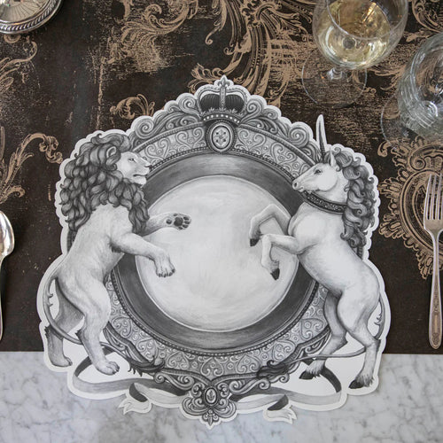 A black and white placemat with a regal crest style illustration featuring a lion on one side and a unicorn on the other resting on a decorative brown table runner with drinking glasses.