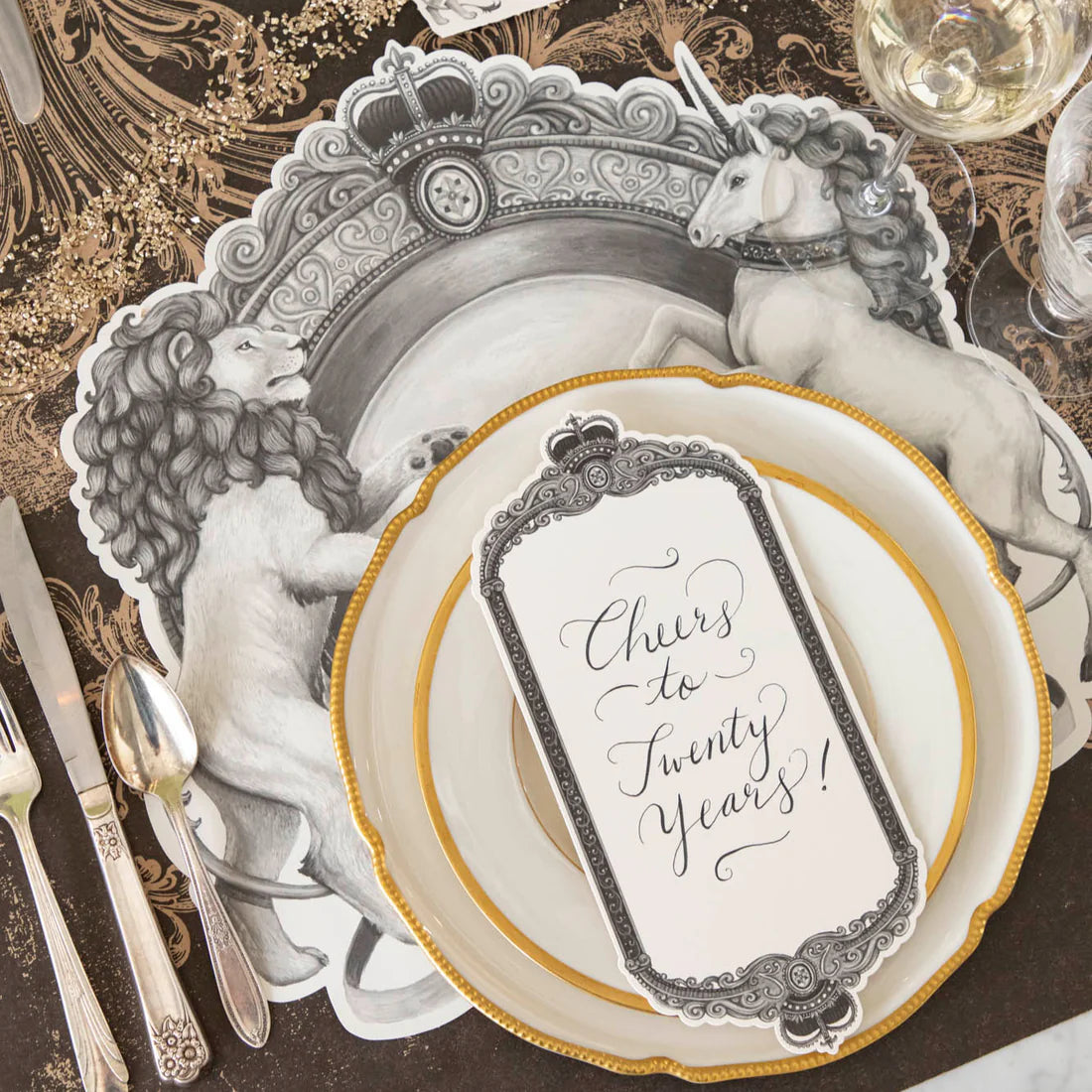 A place setting featuring gold rimmed plates, a decorative brown and beige table runner, a place card, silverware, and a black and white placemat with a regal crest style illustration featuring a lion on one side and a unicorn on the other.
