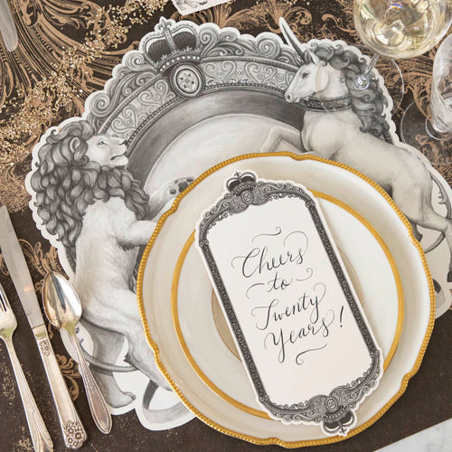 A place setting featuring gold rimmed plates, a decorative brown and beige table runner, a place card, silverware, and a black and white placemat with a regal crest style illustration featuring a lion on one side and a unicorn on the other.