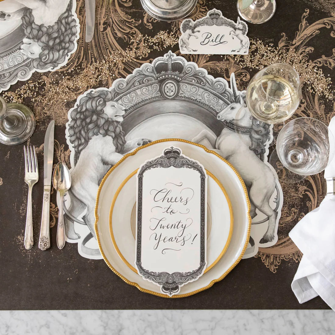 A place setting with a black and white placemat with a regal crest style illustration featuring a lion on one side and a unicorn on the other, with gold rimmed plates, a place card, silverware, and glasses.