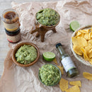 Guacamole in wooden bowls with tortilla chips, limes, and hot sauce on crumpled brown paper.