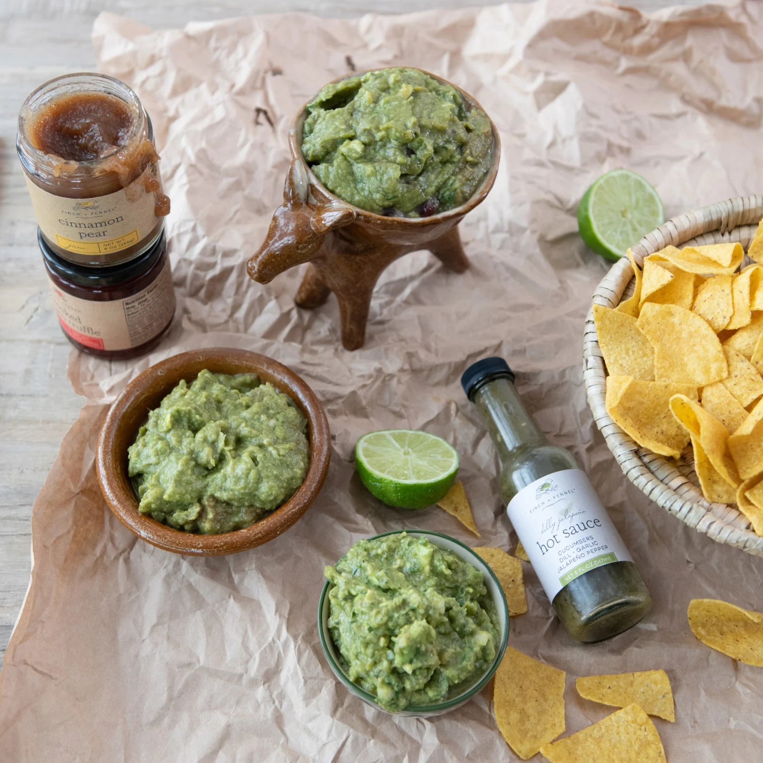 Guacamole in wooden bowls with tortilla chips, limes, and hot sauce on crumpled brown paper.