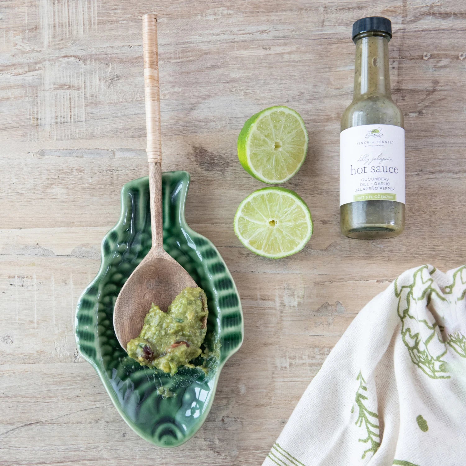 Green dish with a wooden spoon, limes, and hot sauce on a wooden surface