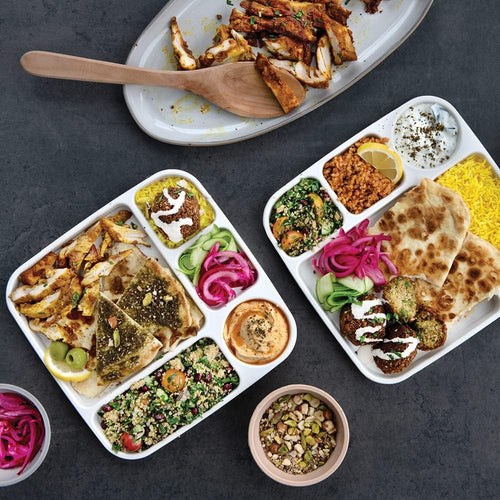 White divided dinner trays full of food with a few bowls of food nearby on a gray surface.