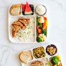 Two compartmentalized lunch trays with a variety of food items including rice, vegetables, and bread on a marble surface.
