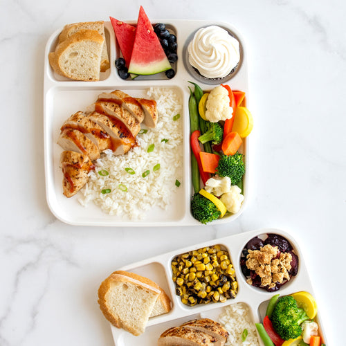 Two compartmentalized lunch trays with a variety of food items including rice, vegetables, and bread on a marble surface.