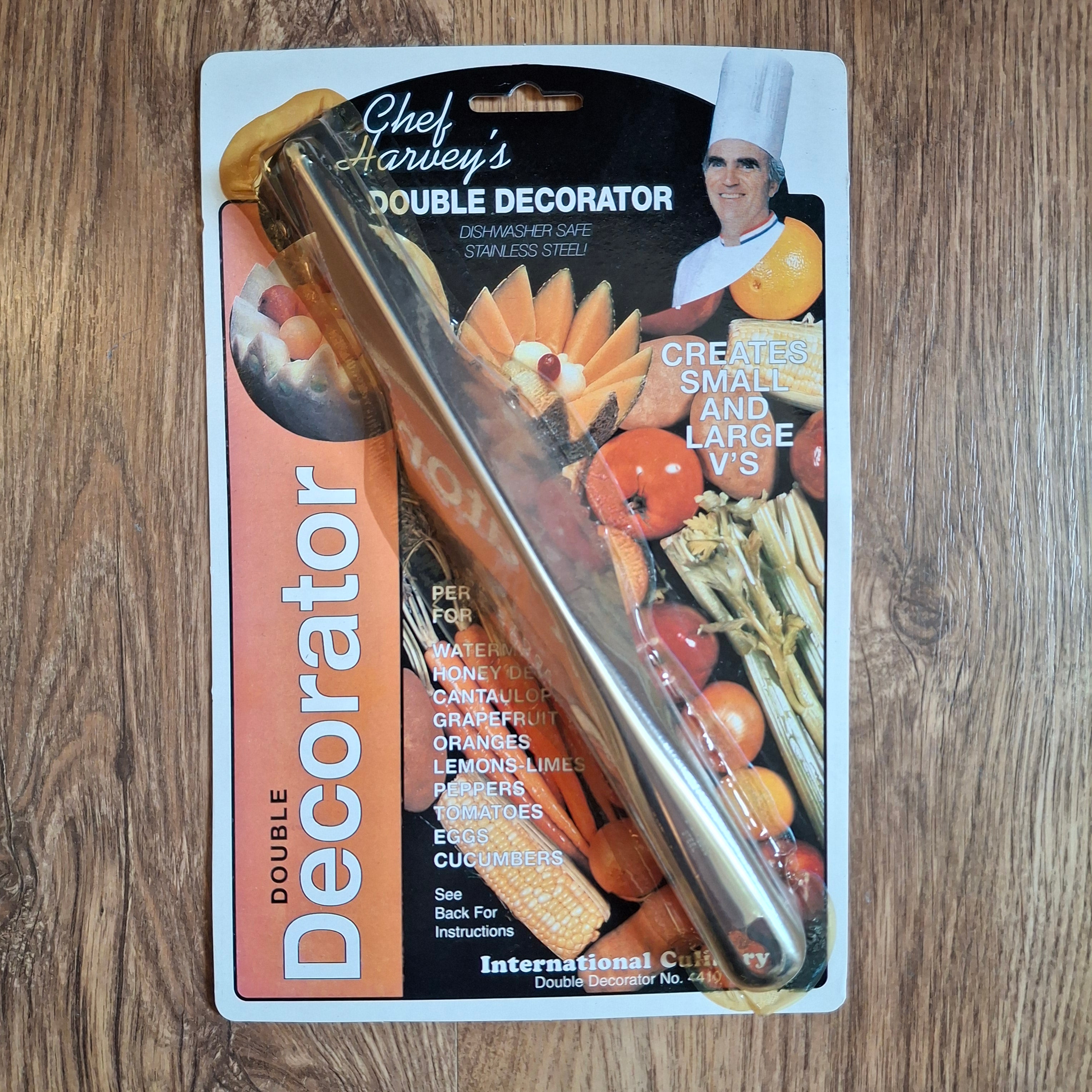 A packaged double-ended stainless steel fruit decorator labeled 'Chef Harley's Double Decorator' on a wood surface. The product is shown in its retail blister packaging with printed graphics of fruits and vegetables and a chef.
