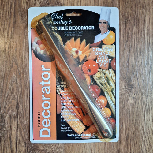A packaged double-ended stainless steel fruit decorator labeled 'Chef Harley's Double Decorator' on a wood surface. The product is shown in its retail blister packaging with printed graphics of fruits and vegetables and a chef.